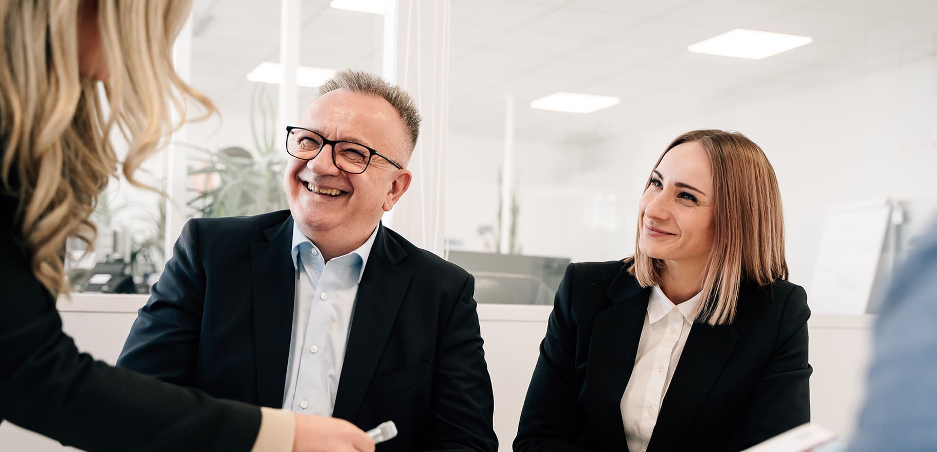 Besprechung eines Produkts im Besprechungsraum zwischen 3 Mitarbeitern die an einem Tisch am sitzen und einer Frau die stehend eine Schraube in der Hand hält.